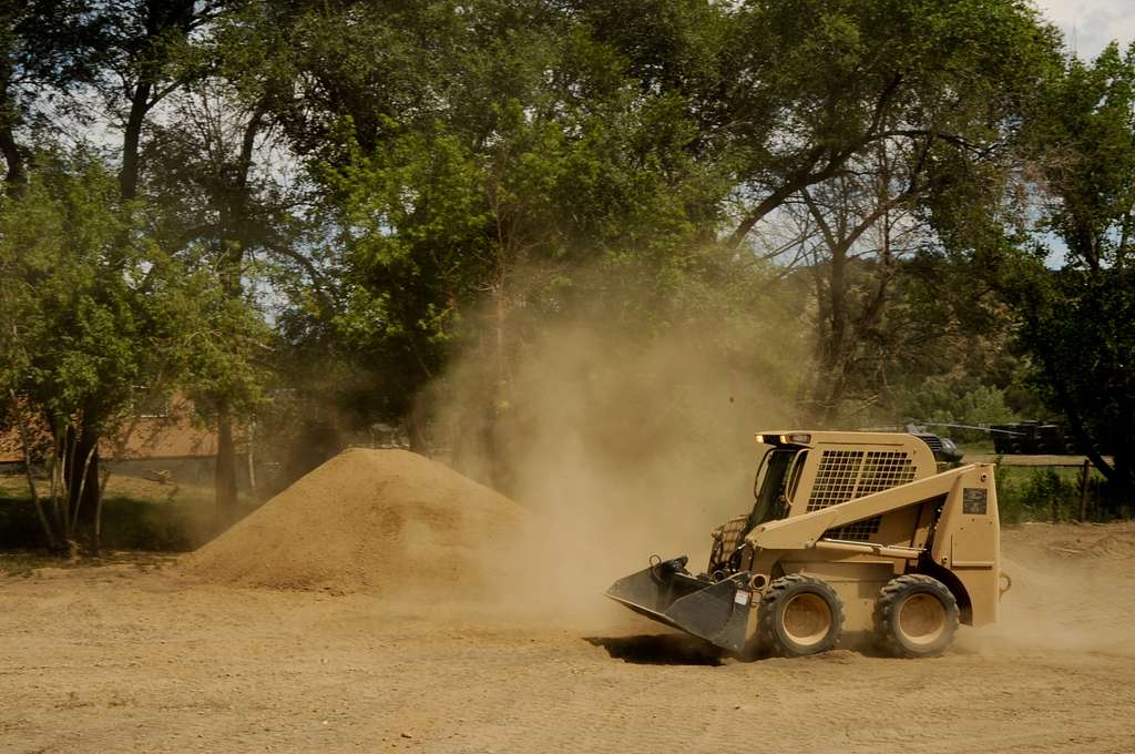 Skid steer loader working in a dusty job site, kicking up sand while moving aggregate — travel drive with frequent stop/restart and dual-wheel independent steering.